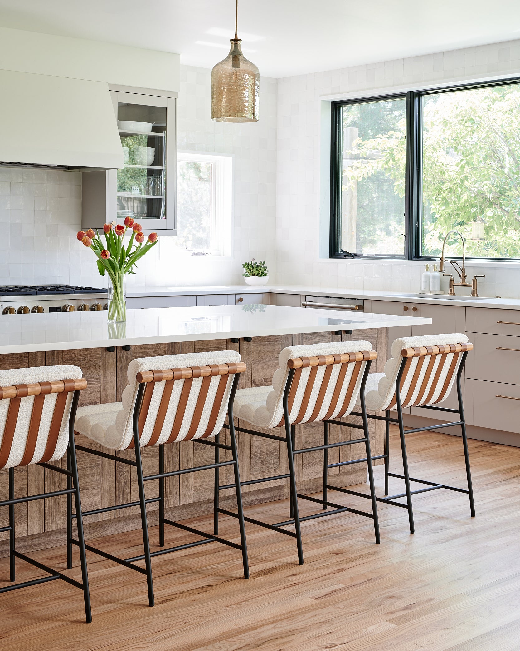 A contemporary kitchen with a large white island countertop, accented by striped barstools with leather and woven fabric. The space includes black-framed windows, white tile backsplash, and warm wooden cabinetry, with a decorative glass pendant light hanging above the island.