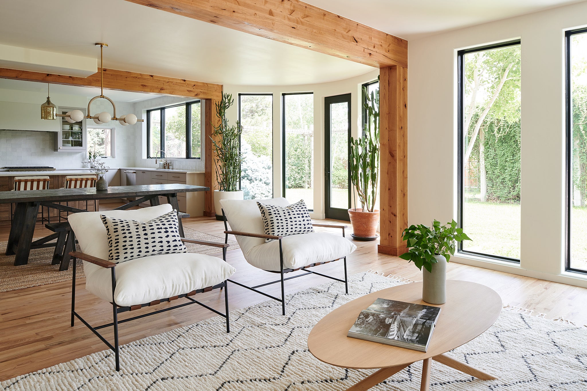 A modern living room featuring two white lounge chairs with wooden arms and black metal frames, positioned on a patterned beige and white rug. The space includes large floor-to-ceiling black-framed windows, wooden beams, and natural decor elements like potted plants and soft lighting.