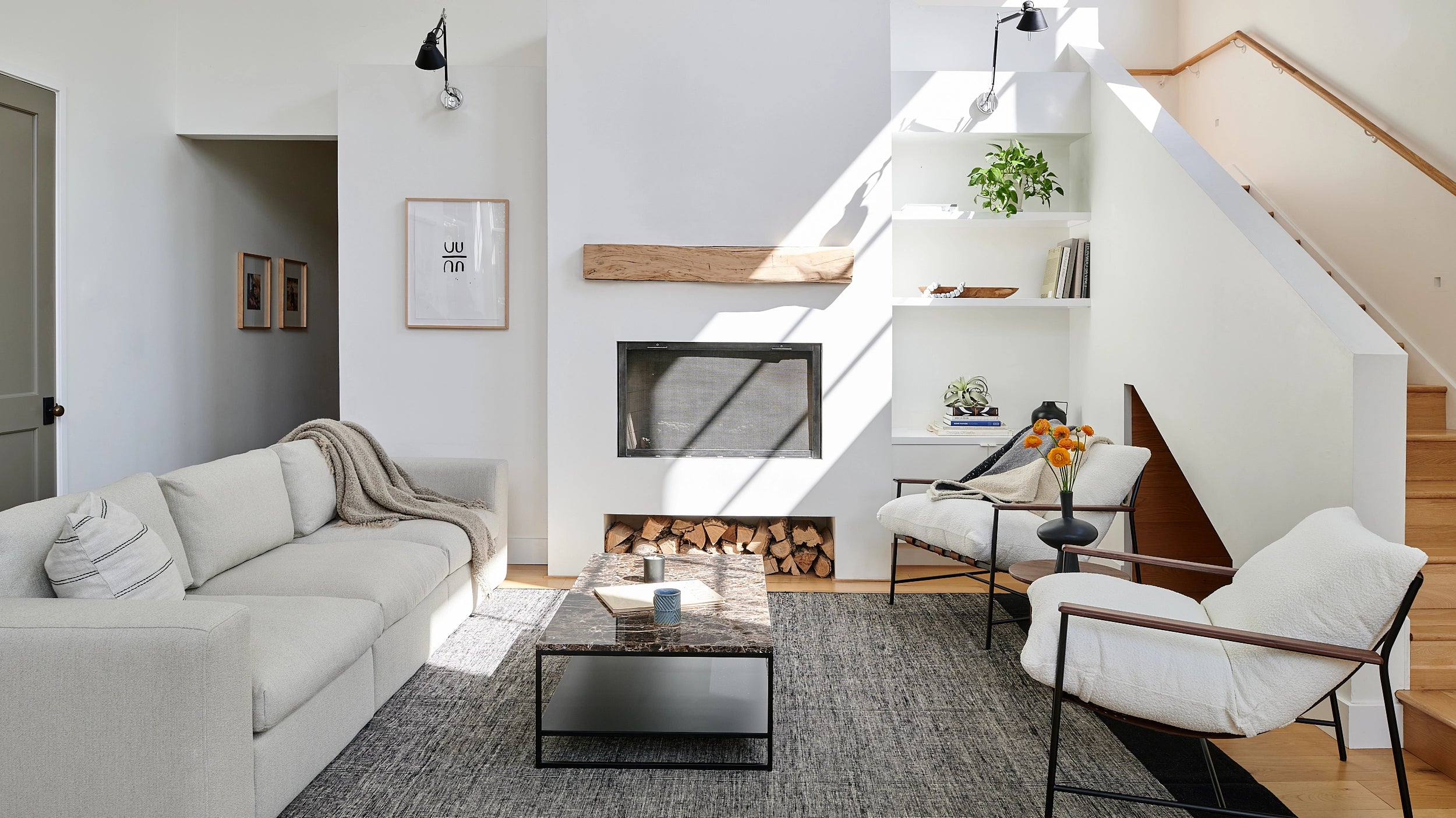 A spacious modern living area featuring a neutral-toned sofa with white cushions, a wooden coffee table, and a patterned rug. The space includes black-framed windows and doors, light wood flooring, and a mix of greenery and natural elements, creating a bright and inviting atmosphere.