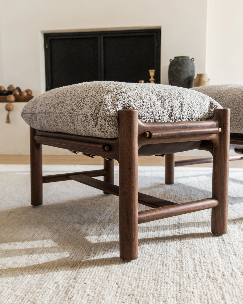 A detailed view of an Avon Ottoman, showcasing its plush light grey cushion and sturdy dark wooden frame. The cushion's textured fabric invites touch, while the wooden frame features elegant cylindrical details that hold the cushion in place.