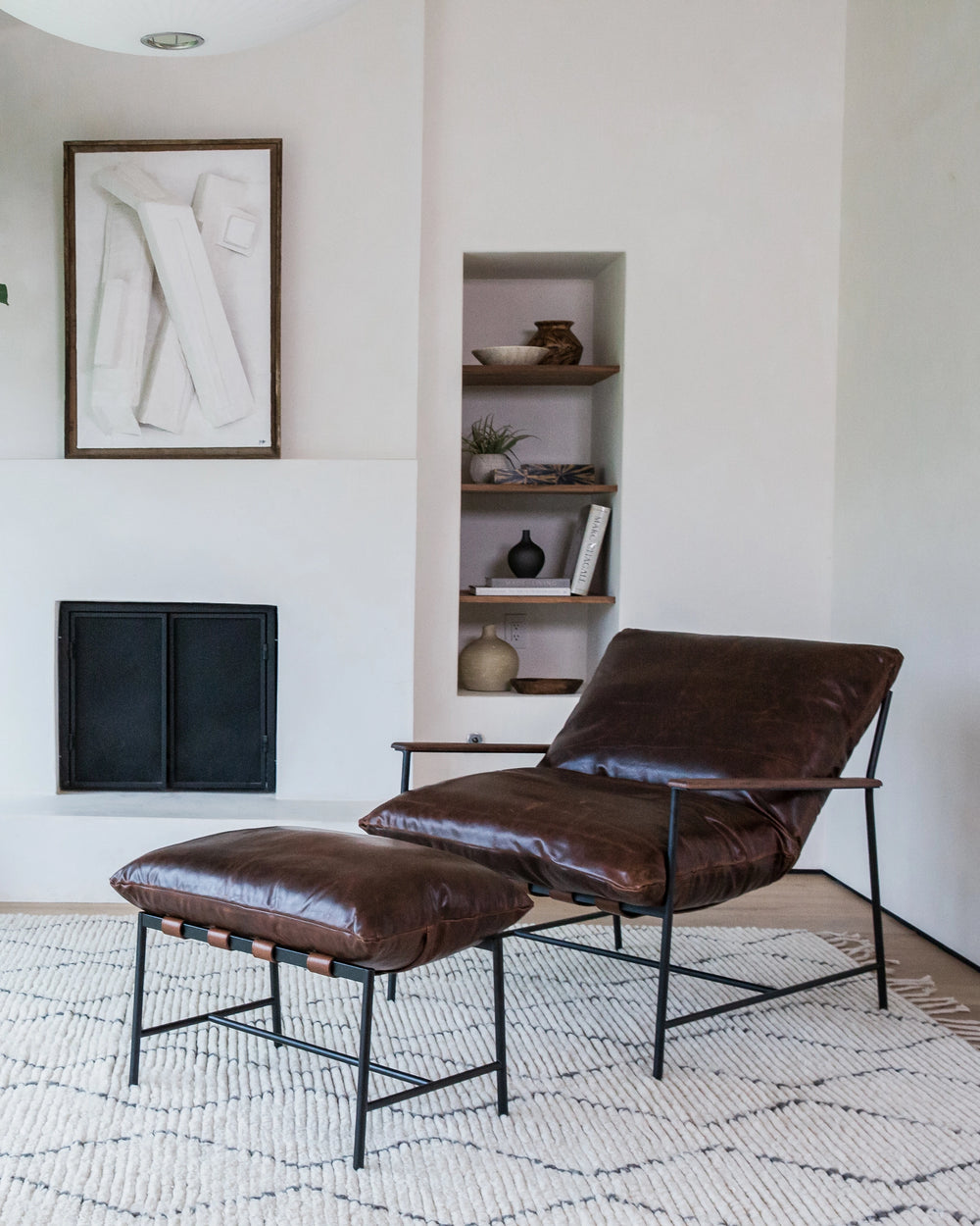 An elegant and serene lounge space featuring a brown leather Vail Lounge Chair with a matching ottoman. Both the chair and ottoman have black metal frames and are set upon a textured off-white area rug. 